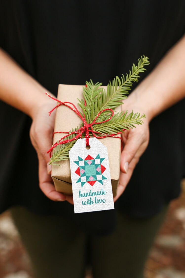 A person holding a package wrapped in brown wrapping paper and tied with a piece of red twine with a piece of greenery and a gift tag attached that says, \"Handmade with Love\"