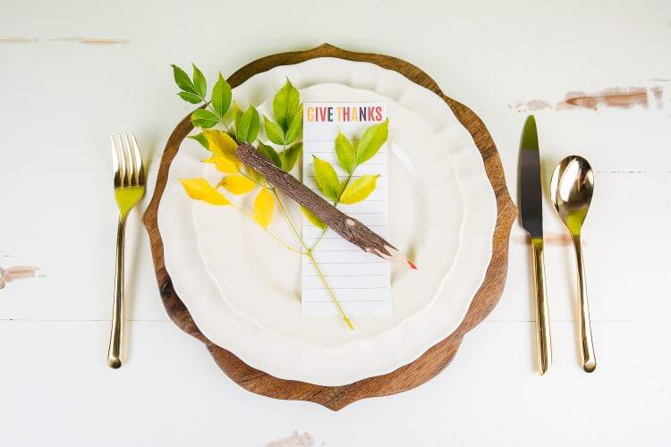 A place setting of a plate, cutlery and some greenery, a wooden pencil and a piece of paper that says, \"Give Thanks\" sitting on top of the plate