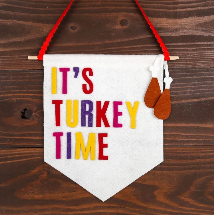 A white banner hanging from a red piece of rick rack and the banner says, \"It\'s Turkey Time.  The banner has two felt turkey drumsticks hanging from it.