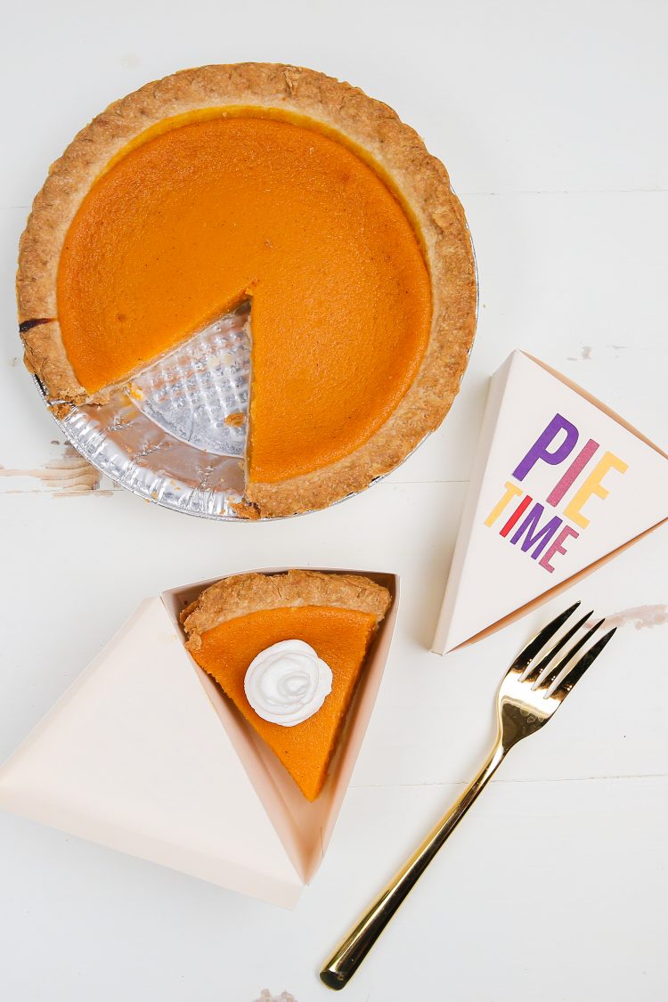 A pumpkin pie sitting next to a fork and two boxes, one with a piece of pie in it and the other one closed with the saying \"Pie Time\" on it