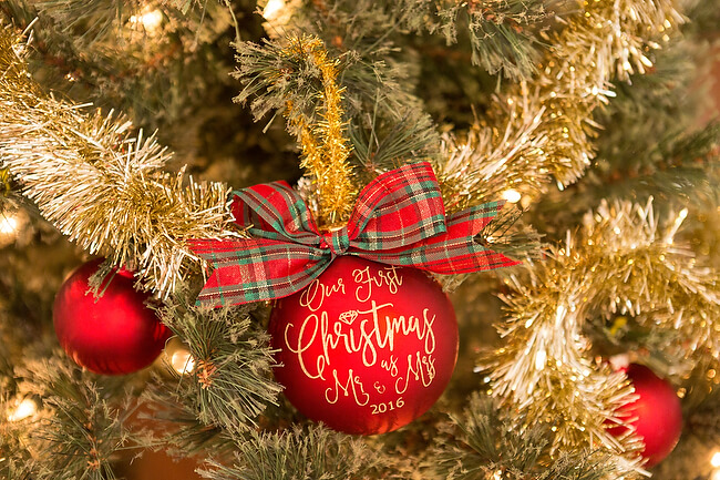 A red ornament hanging from a Christmas tree and the ornament is decorated with green and red paid ribbon and says, \"Our First Christmas Mr. & Mrs. 2016\"