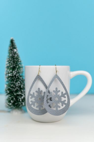 Snowflake design earrings hanging from a coffee cup that is sitting on a table next to a small fake tree