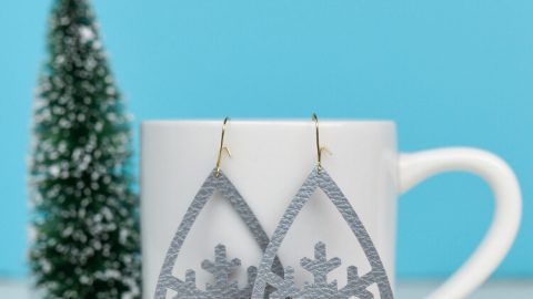 Snowflake design earrings hanging from a coffee cup that is sitting on a table next to a small fake tree