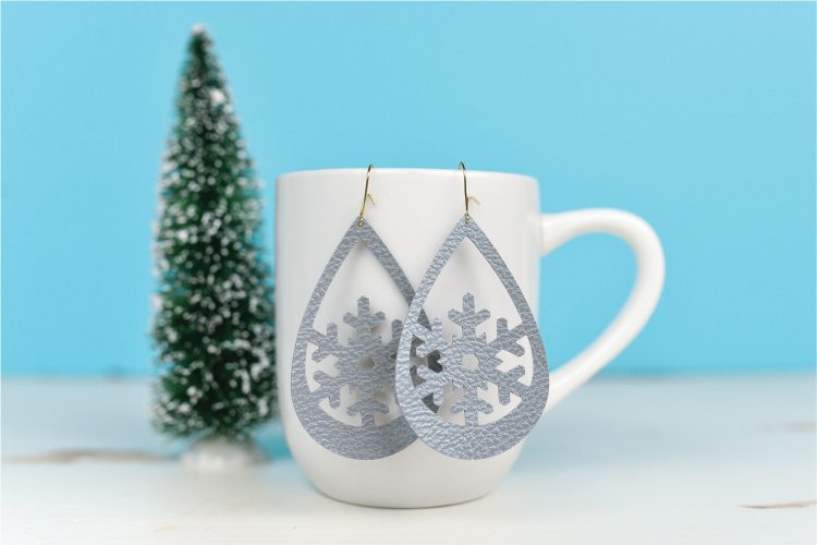 Close up of snowflake design earrings hanging from a coffee cup that is sitting on a table next to a small fake tree