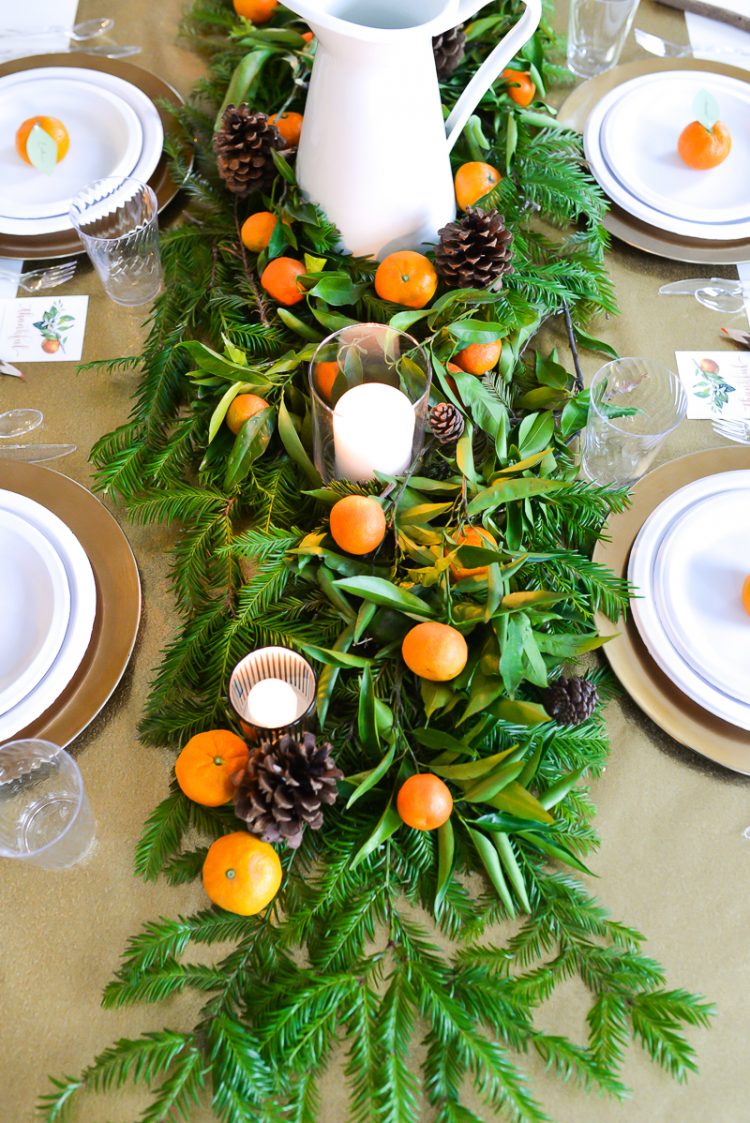 A table topped with a table runner decorated with greenery and candles and oranges along with place settings of plates, cutlery and water glasses