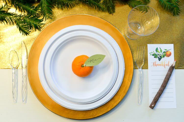 A place setting of a water glass, cutlery and a plate with an orange on it all next to a pen sitting on top of a piece of paper with the word \"Thankful\" on it