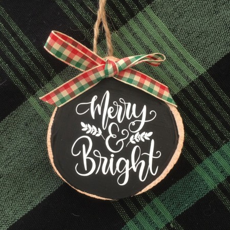 A round wooden ornament lying on a green plaid tablecloth, and it says, "Merry & Bright"