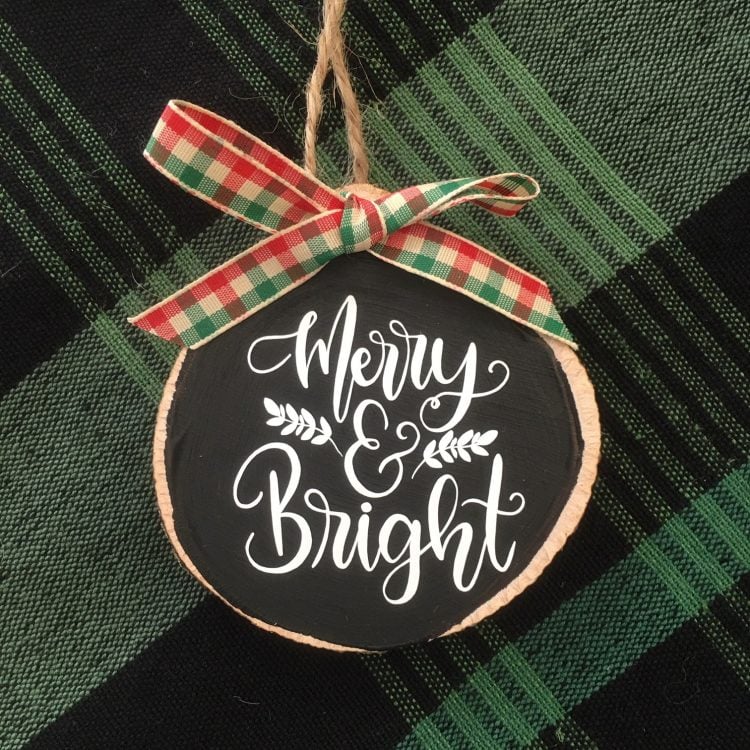 A round wooden ornament lying on a green plaid tablecloth, and it says, \"Merry & Bright\"