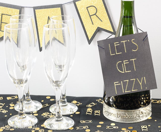 A table with champagne flutes and a bottle of champagne that has a sign on it that says, \"Let\'s Get Fizzy\"