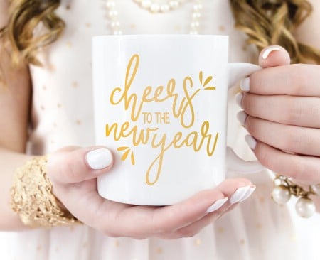 Woman holding a white coffee mug with the saying Cheers to the New Year in gold lettering