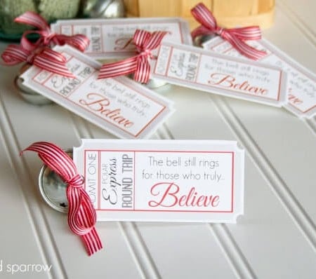 Close up of Christmas gift tags with a red bow and a jingle bell tied to each and they say, "Admit One - Polar Express Round Trip - The bell still rings for those who truly believe"