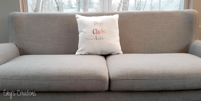 A living room with a large window and a pillow sitting on a couch and the pillow says, \"Pop Clink Kiss\"