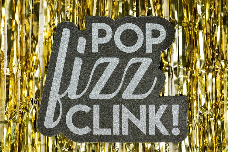 A close up of a sign that says, \"Pop Fizz Clink!\" hanging from a background of gold tinsel
