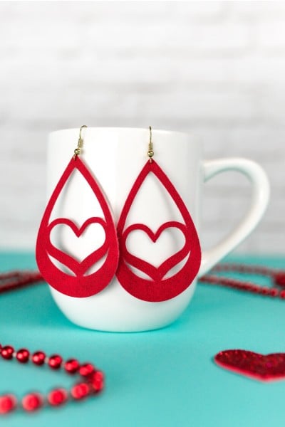 Valentine suede earrings hanging from a coffee cup that is sitting on a table with red beaded necklace on it