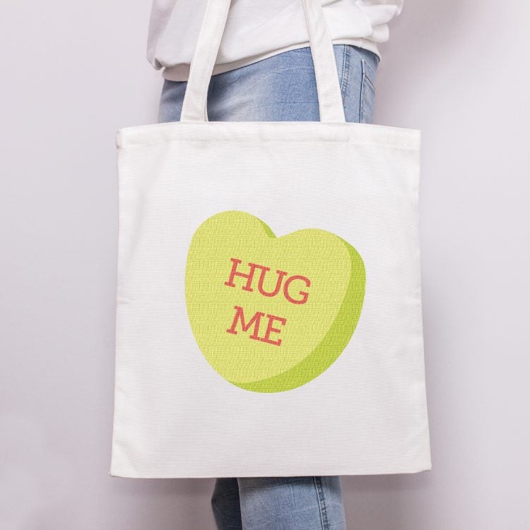 A person holding a white tote bag that is decorated with a green Valentine conversation heart that says, \"Hug Me\"