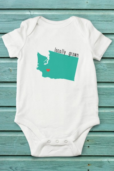 A white onesie laying on an aqua blue table, and it is decorated with an image of the state of Washington and says, "Locally Grown"