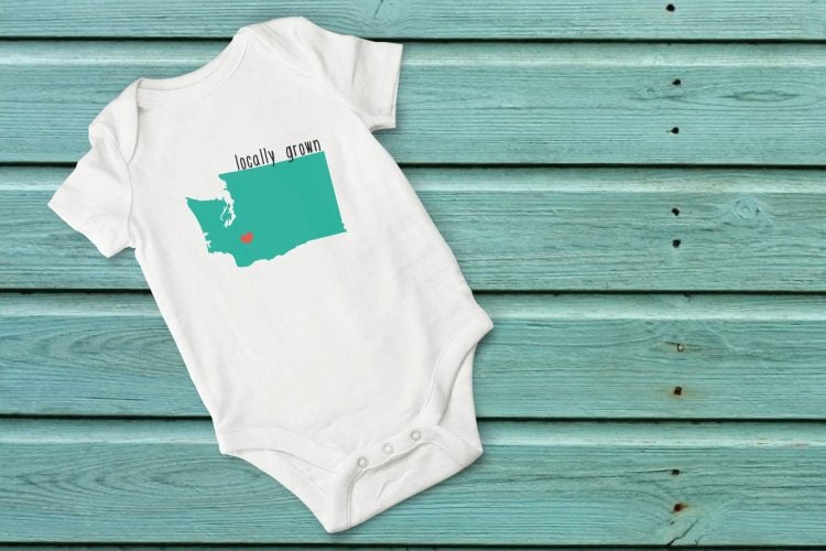 A white onesie laying on an aqua blue table, and it is decorated with an image of the state of Washington and says, \"Locally Grown\"