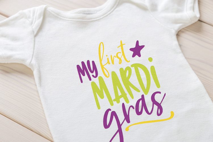 Close up of a white shirt laying on a table, with the saying, \"My First Mardi Grads\" on it