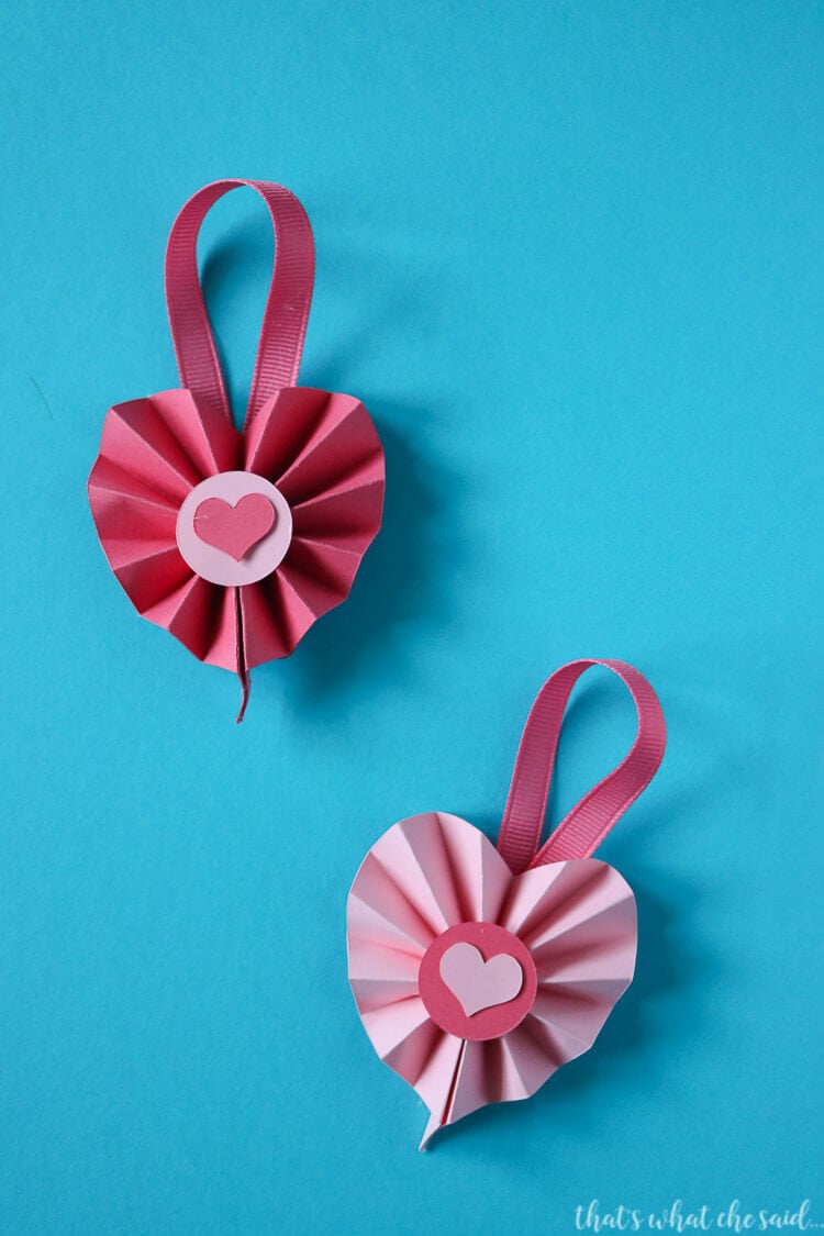 Two pieces of accordion folded paper that are in the shape of hearts with a pink dot on top of each one and a small pink heart on top of the dot on each one and ribbon attached to each for hanging them