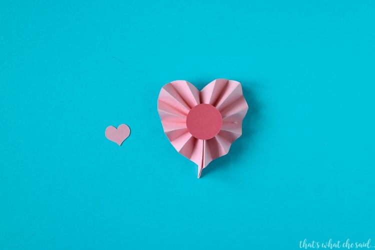 A piece of accordion folded paper that is in the shape of a heart with a pink dot on top of it and a small pink heart next to it
