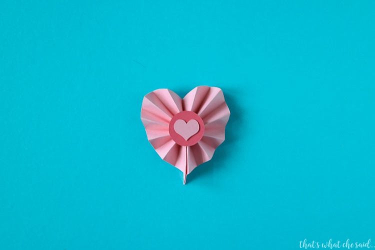 A piece of accordion folded paper that is in the shape of a heart with a pink dot on top of it and a small pink heart on top of the dot