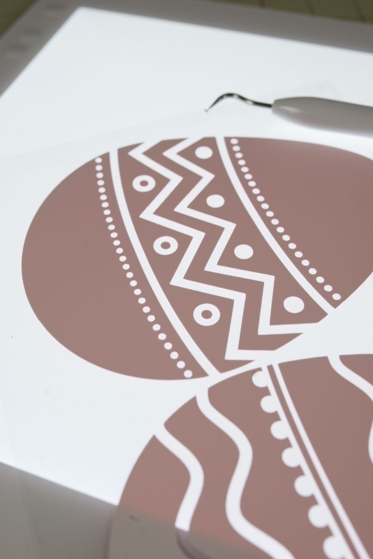 Close up of two brown colored paper cut Easter eggs decorated with white iron-on vinyl and a Cricut weeding tool