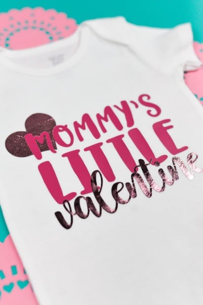 A white onesie with a heart and quote "Mommy's Litte Valentine".  Onesie is surrounded by pink doilies.