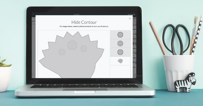 A screen shot of the Hide Contour feature in Cricut Design Space on an open laptop sitting on top of a table along with a plant and a glass that contain a pencil, a paintbrush and a scissors