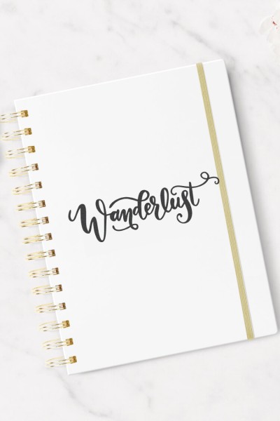 A white journal sitting on a table, decorated with the word, "Wanderlust"