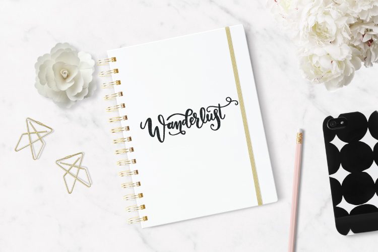 Close up of a pencil, paperclips, white flowers and a white journal sitting on a table, decorated with the word, \"Wanderlust\"