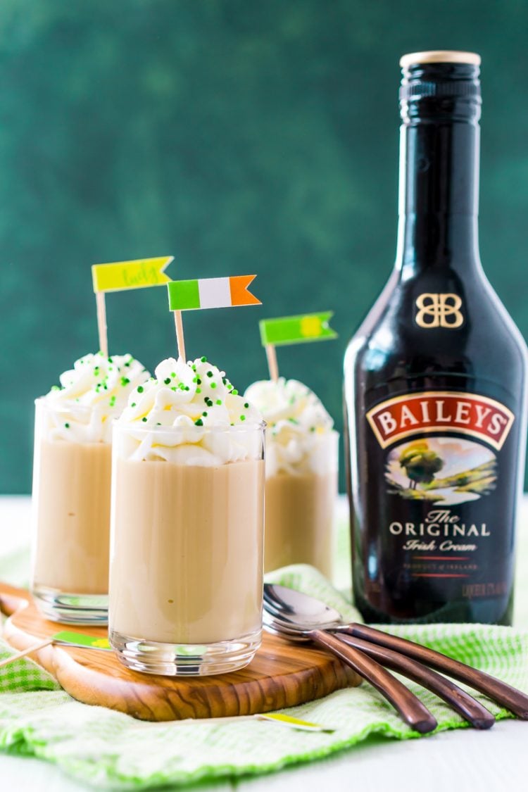 A bottle of Bailey\'s Irish Creme on a table along with three spoons and three decorated beverages