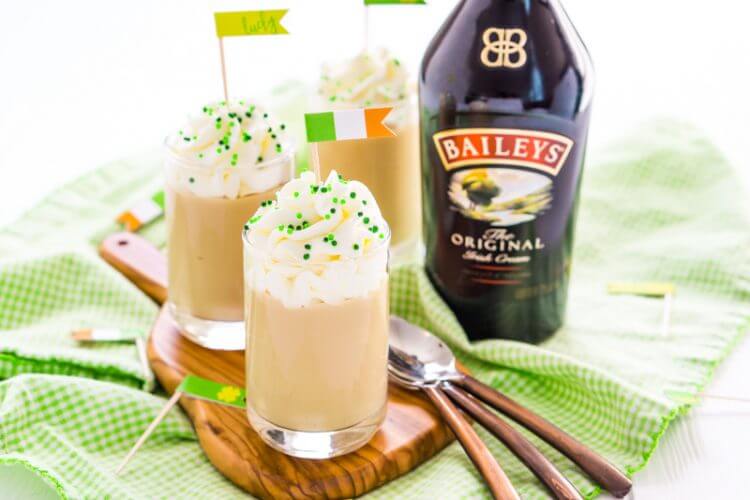 A close up of a bottle of Bailey\'s Irish Creme on a table along with three spoons and three decorated beverages
