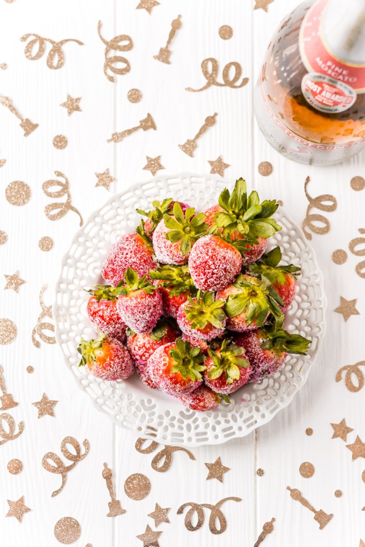 Gold glittery images of stars, dots swirls and Oscar award statues confetti on top of a table that holds a bowl of sugar covered strawberries and a bottle of wine