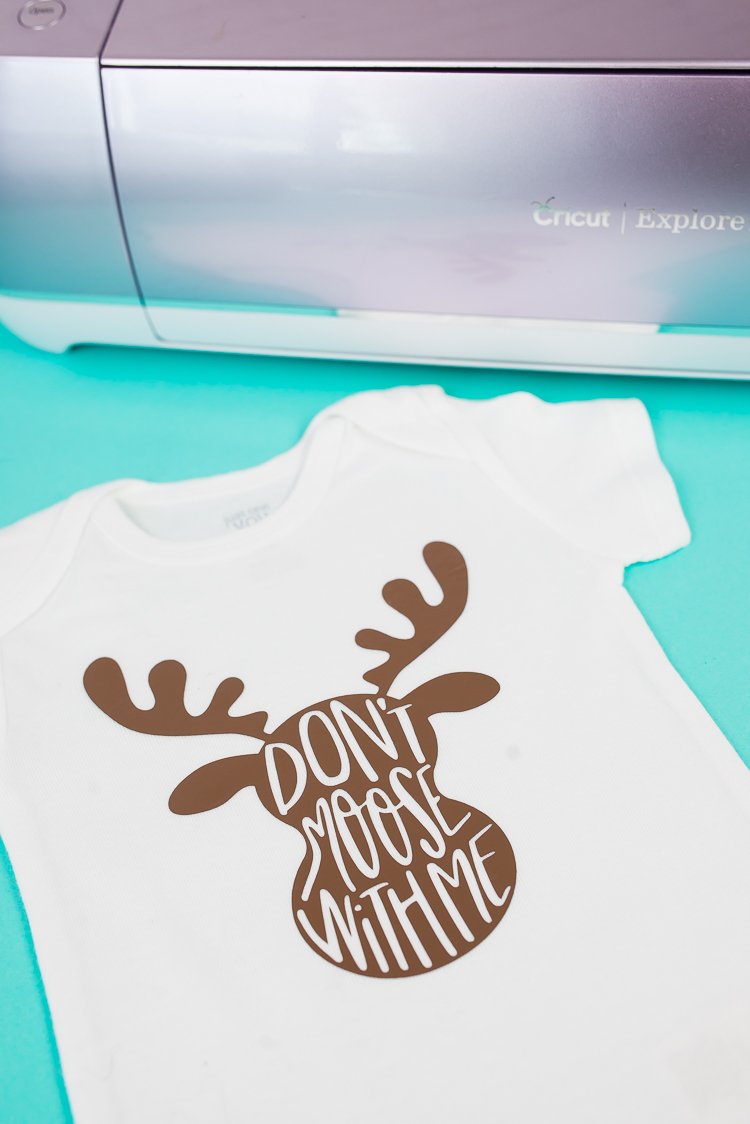 A white onesie that has an image on a moose on it and the saying, \"Don\'t Moose with Me\" next to a Cricut Explore 2 machine