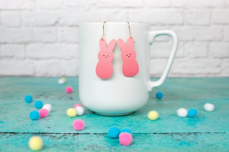 A white coffee mug sitting on an aqua blue table with colorful pompoms scattered around it and two pink bunny earrings hanging from the mug.