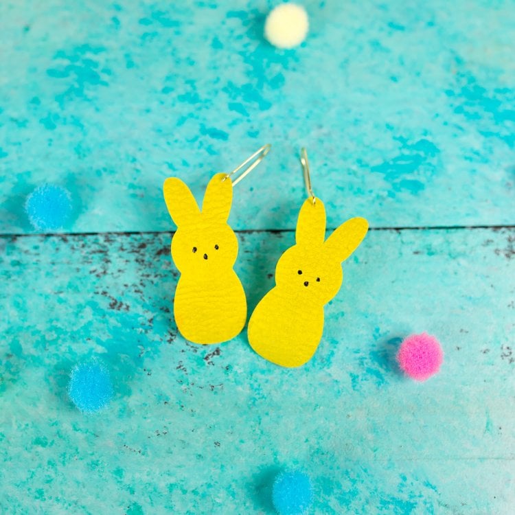 Colorful pompoms scattered all around two yellow bunny earrings and all sitting on an aqua blue table