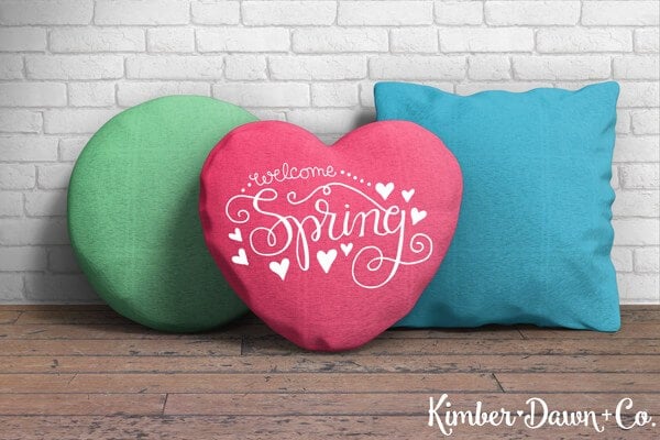 A round green pillow, a blue square pillow and a red heart shaped pillow that says, \"Welcome Spring\" all sitting on a wooden bench up against a white brick wall