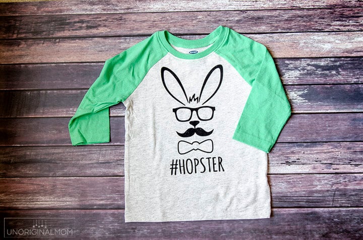 A baseball style shirt lying on top of a wooden table and the shirt has a design on it of an Easter bunny with a mustache and bowtie and says, \"#Hopster\"