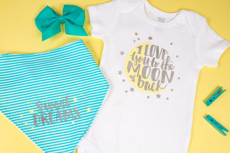 A baby girl aqua colored bow next to a white onesie that says, \"I Love You to the Moon and Back\" and a baby bib that says, \"Sweet Dreams\"