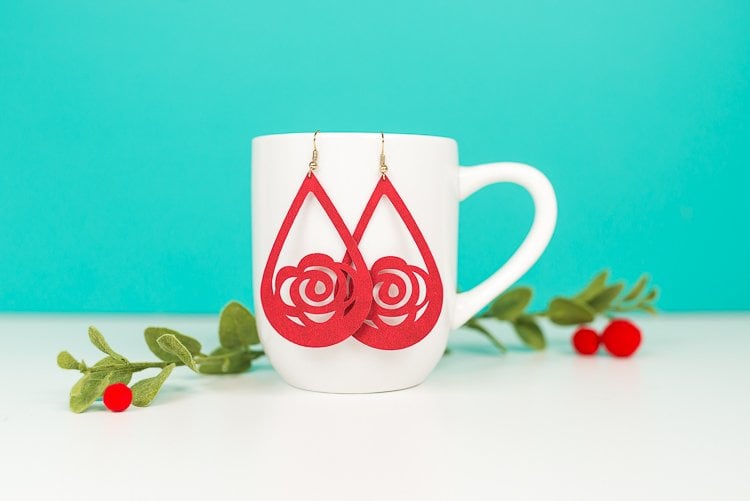 Some greenery next to a coffee cup that has two suede flower earrings hanging from it