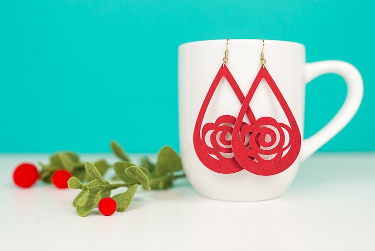 A close up of some greenery next to a coffee cup that has two suede flower earrings hanging from it