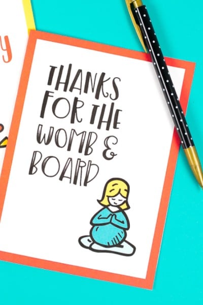 A pen sitting on top of a greeting card that has an image of a pregnant woman on it and says, "Thanks for the Womb and Board"