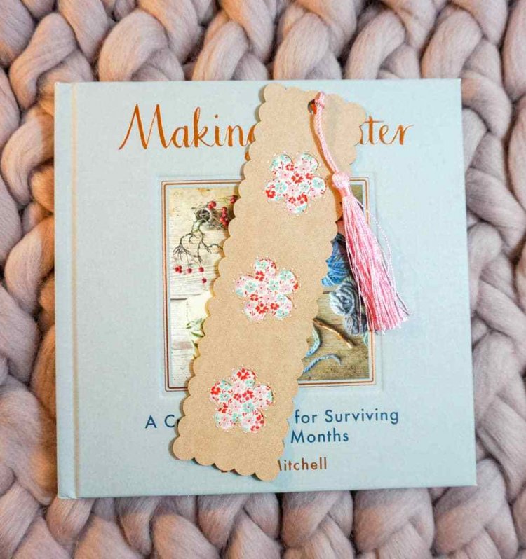 A book with a bookmark laying on top of it that has a tassel attached to it and the bookmark is decorated with a floral design