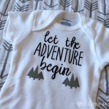 A white onesie laying on a blanket with arrows on it and the onesie is decorated with trees and the saying, "Let the Adventure Begin"