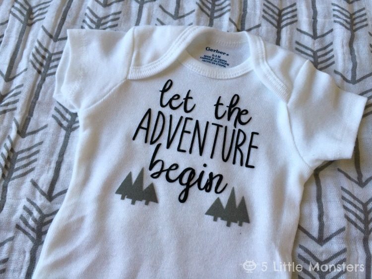 A white onesie laying on a blanket with arrows on it and the onesie is decorated with trees and the saying, \"Let the Adventure Begin\"