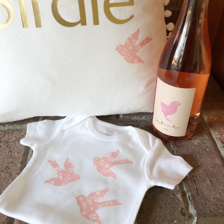A bottle of wine, a pillow next to a onesie that is decorated with images of birds