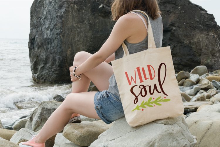 A woman sitting on a rock with a canvas bag hanging over her shoulder that says, \"Wild Soul\"