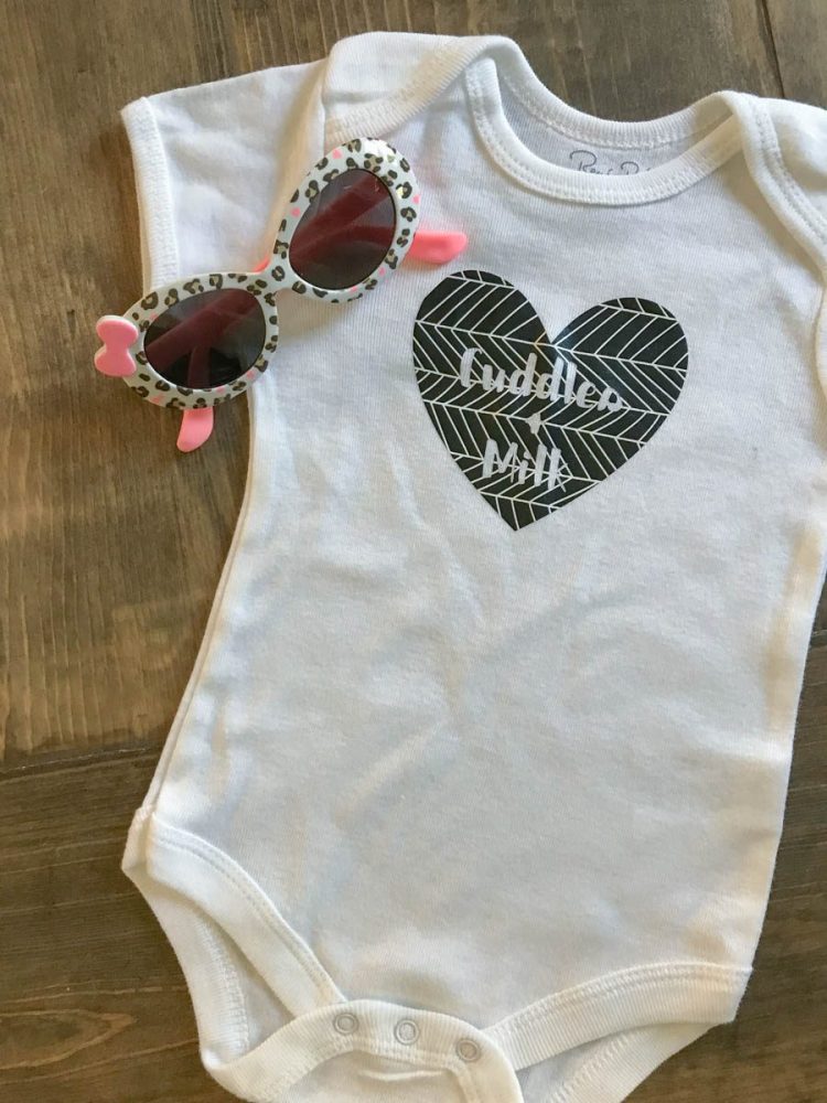 A pair of little baby sunglasses and a white onesie decorated with a heart and the saying, \"Cuddles & Milk\"