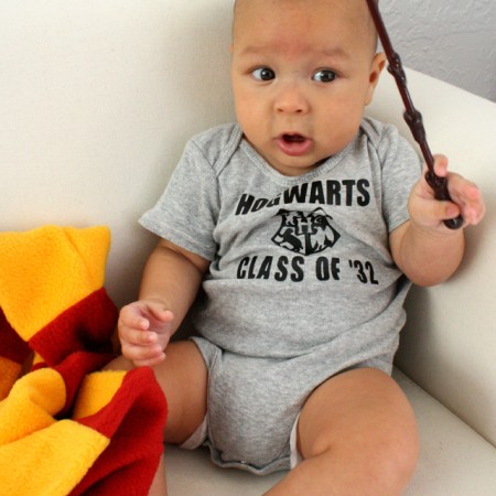 A little boy sitting on a white couch while holding a brown stick and a red and yellow blanket partially covering his leg.  He is wearing a gray onesie that says, "Hogwarts Class of "32"
