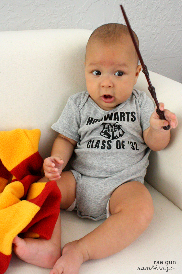 A little boy sitting on a white couch while holding a brown stick and a red and yellow blanket partially covering his leg.  He is wearing a gray onesie that says, \"Hogwarts Class of \"32\"